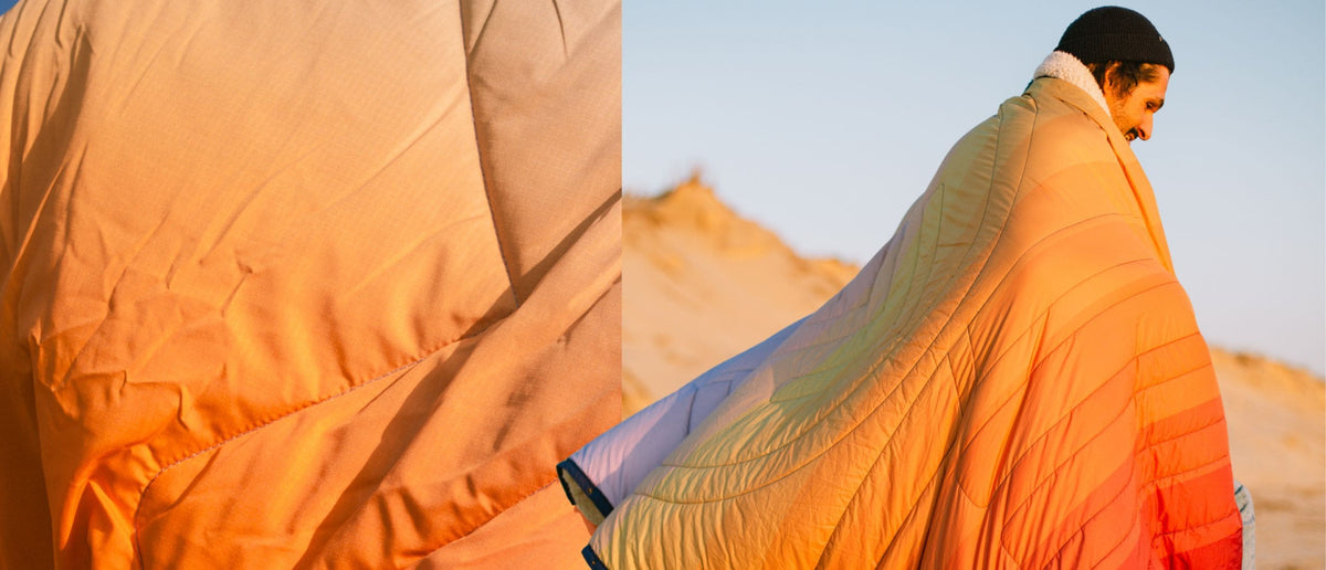 A quilted puffer blanket with a soft white sherpa collar, shown in a close-up detail view on the left and worn by a person in a desert setting on the right. The blanket features a vibrant gradient pattern transitioning from orange and yellow to light blue and purple. CloudTouch® Blanket
