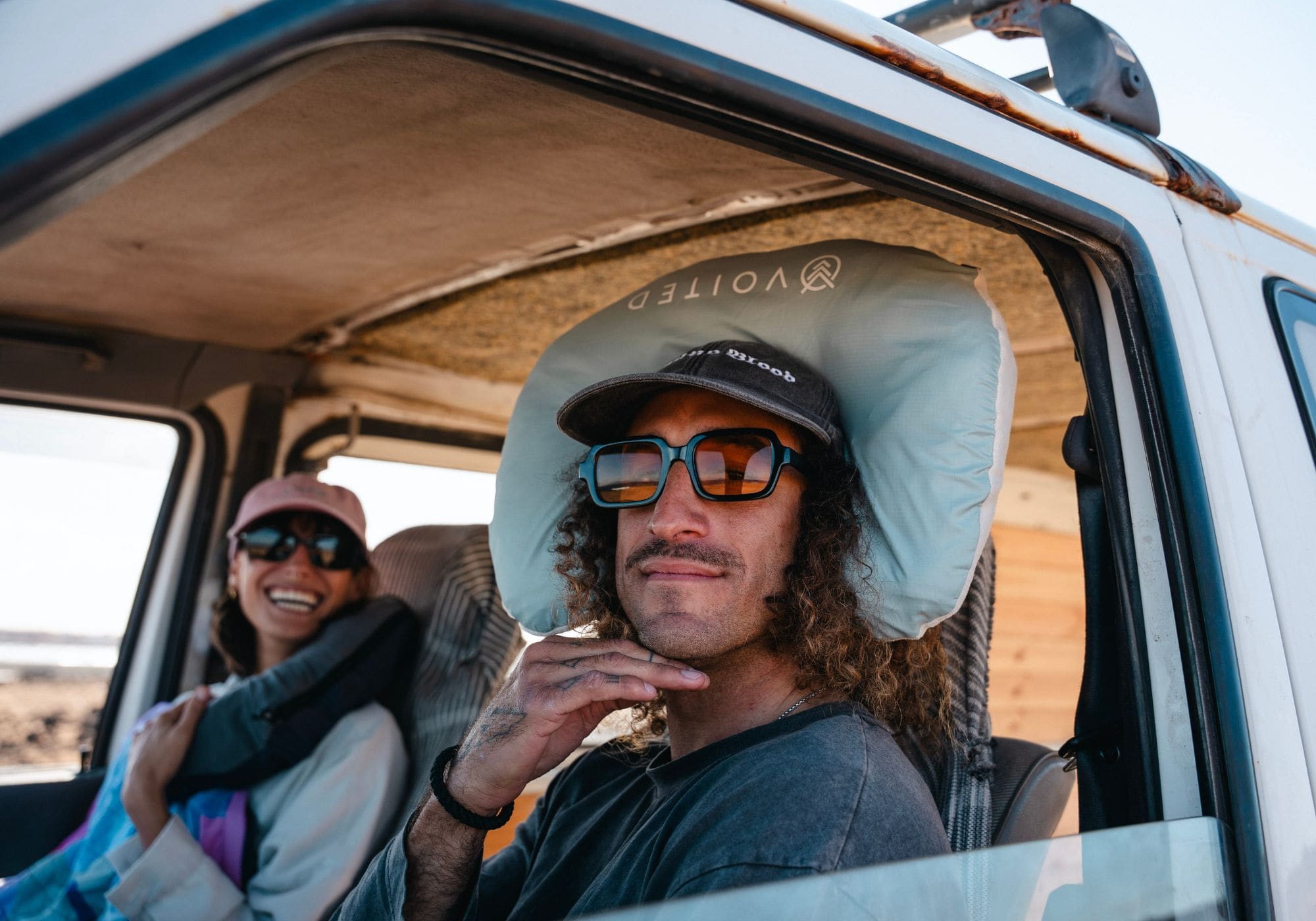 A man sitting in the driver's seat of a vehicle wearing a light blue Voited NeckPackTravel Blanket VOITED under his cap, with a woman smiling in the background. 