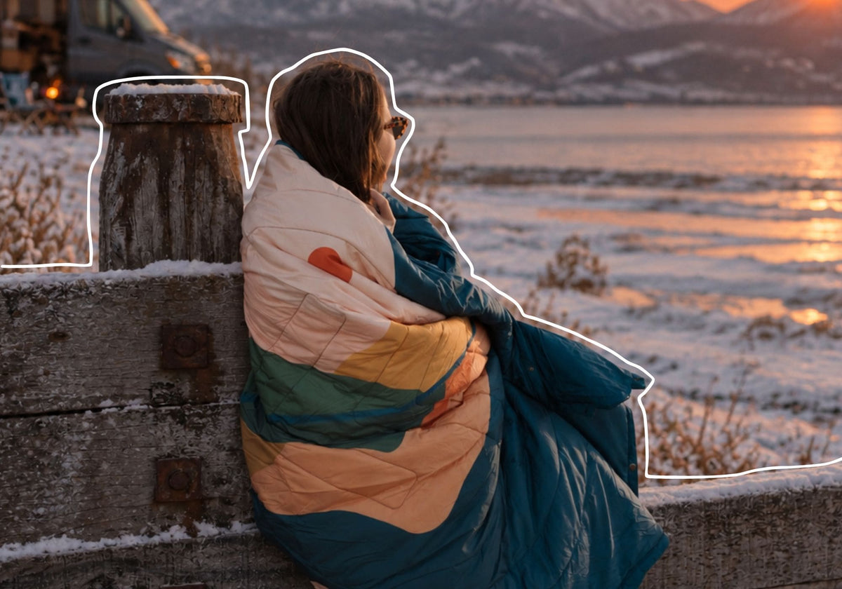 A person sitting outdoors by a snowy lakeside at sunset, wrapped in a VOITED Cloudtouch blanket with horizontal stripes of cream, mustard yellow, teal green, and dark blue. The background shows snow, water, and mountains during golden hour. AI modified